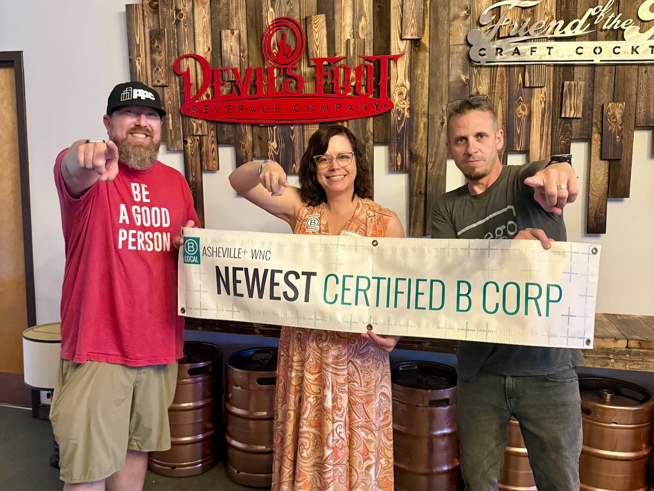 Three people holding the B Local Asheville and WNC newest certified B Corp banner in front of the Devil’s Foot Beverage Company sign